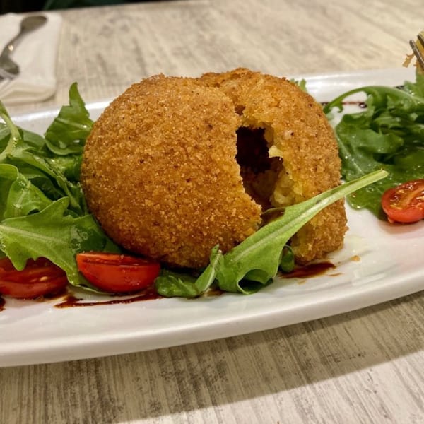 A large, golden-brown arancini with a bite taken out, served on a bed of arugula and cherry tomatoes.
