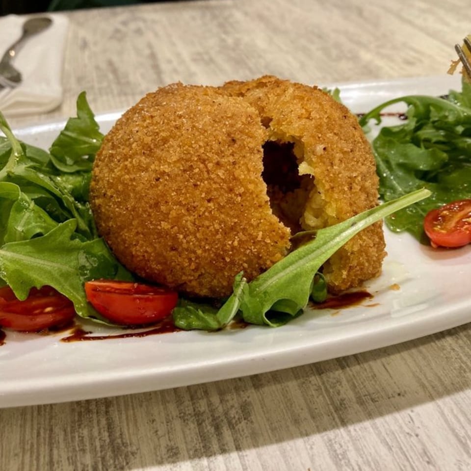 A large, golden-brown arancini with a bite taken out, served on a bed of arugula and cherry tomatoes.
