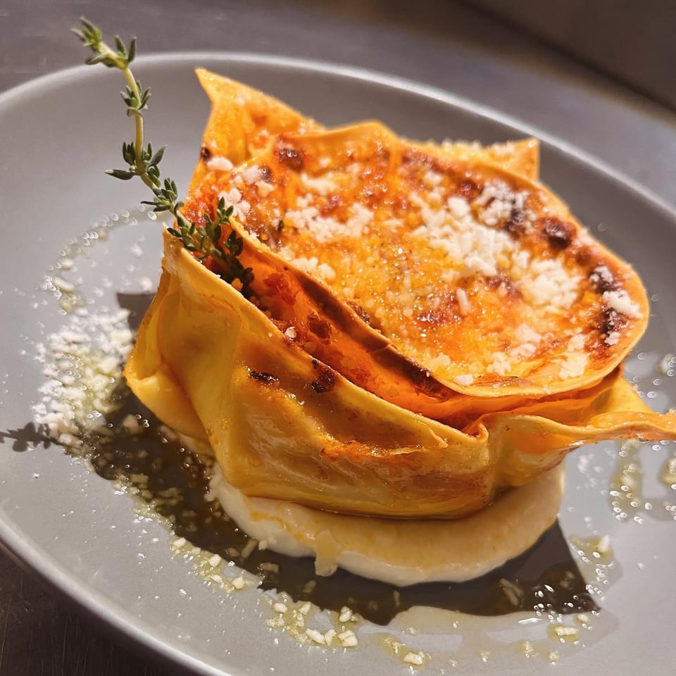 From Scratch's Italian lasagna, topped with parmesan cheese and a sprig of thyme, on a gray plate.
