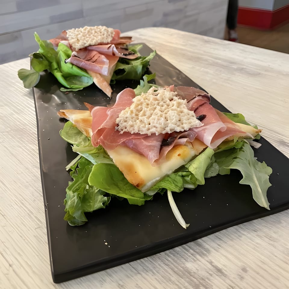 Two Italian flatbreads with prosciutto, cheese, lettuce, and a white topping on a black platter.
