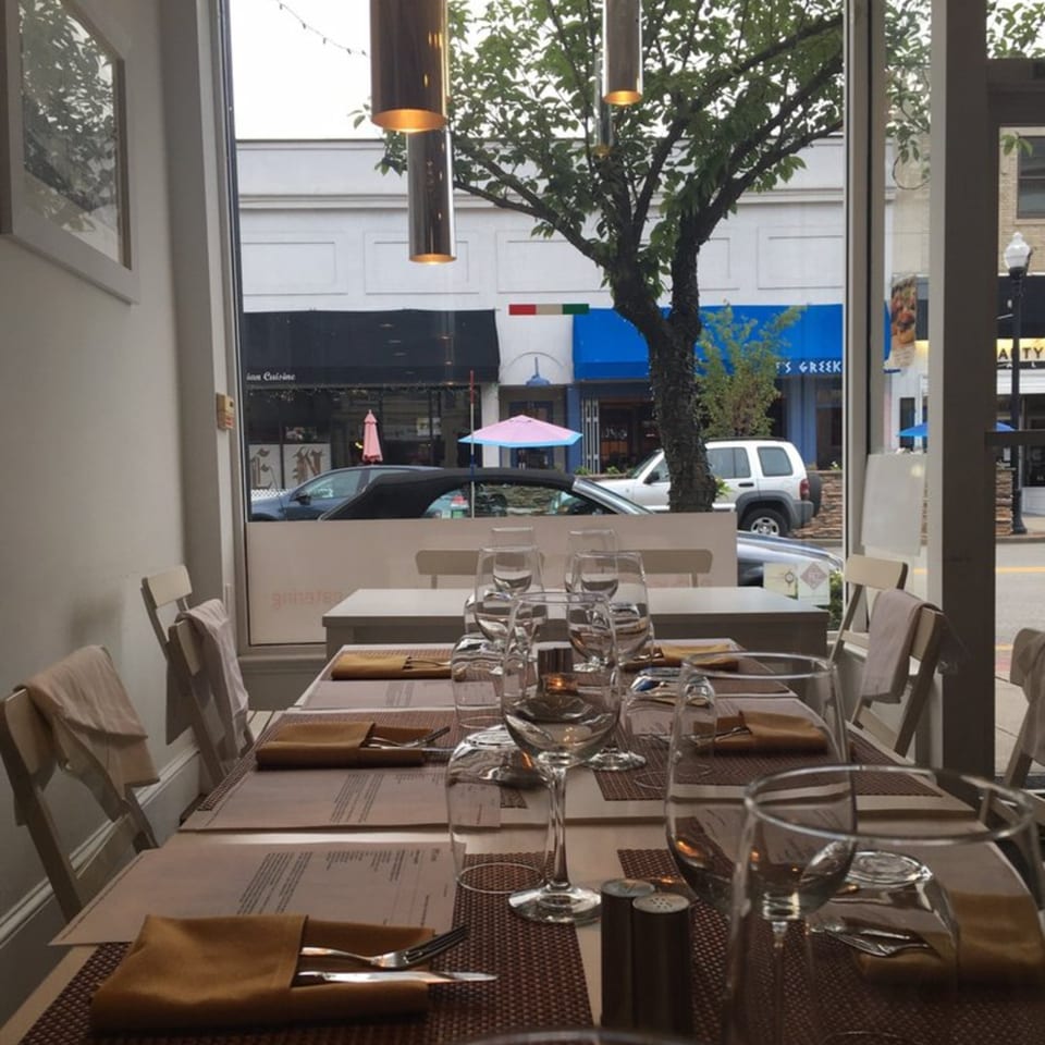 Set tables with wine glasses and menus, looking out a window onto a street with trees and buildings.