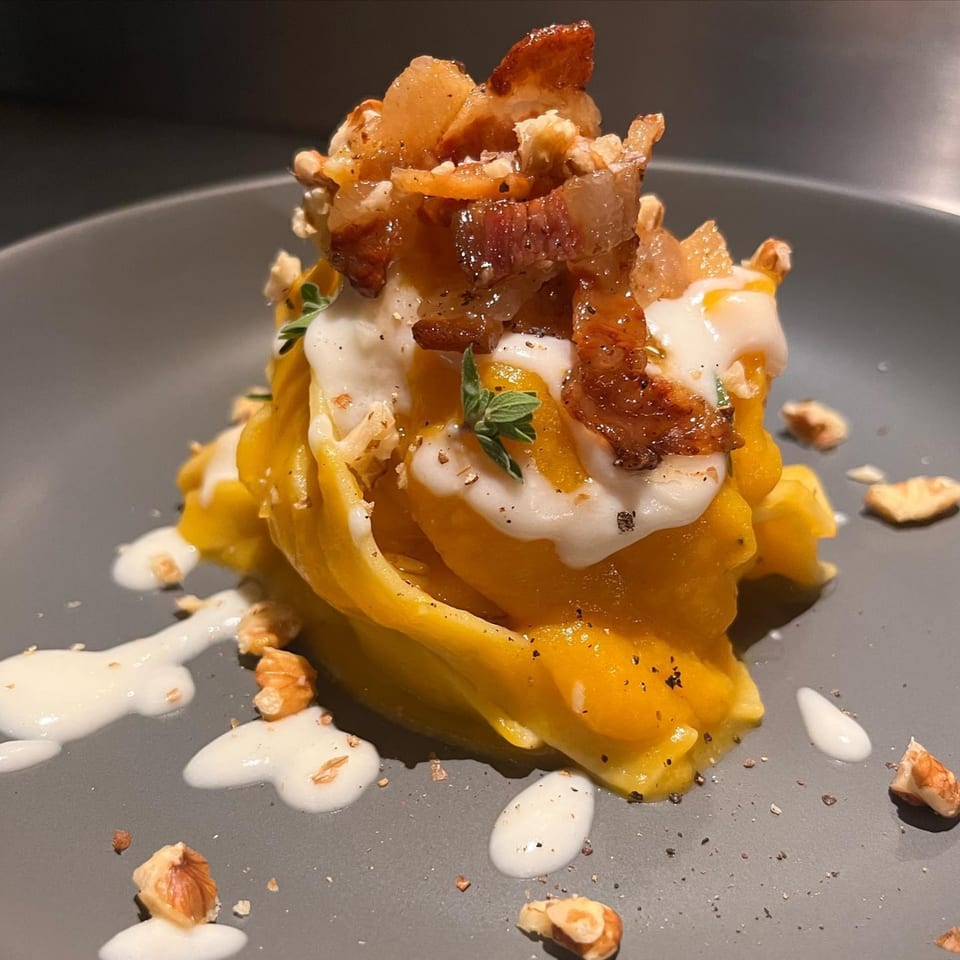 From Scratch's Italian dish with yellow pasta, white sauce, bacon, and walnuts on a gray plate.