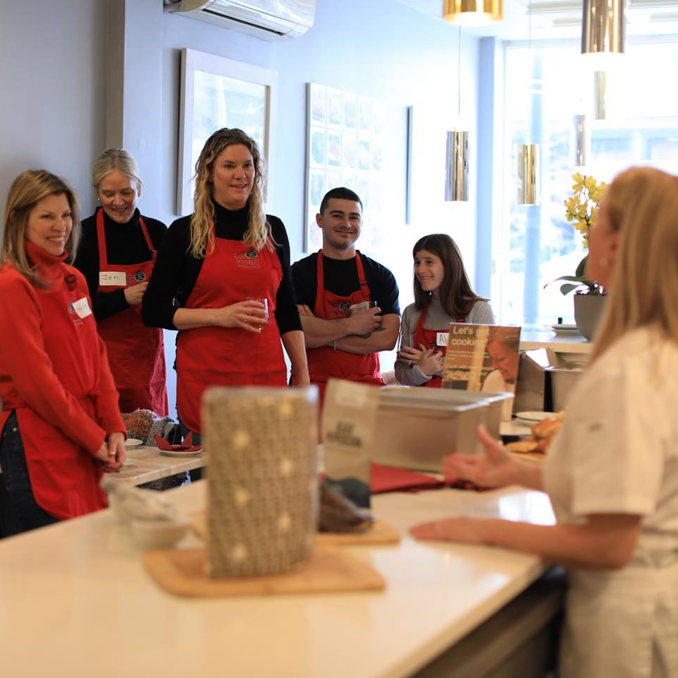 Five people in red aprons listen to a chef in a bright, modern kitchen at From Scratch.