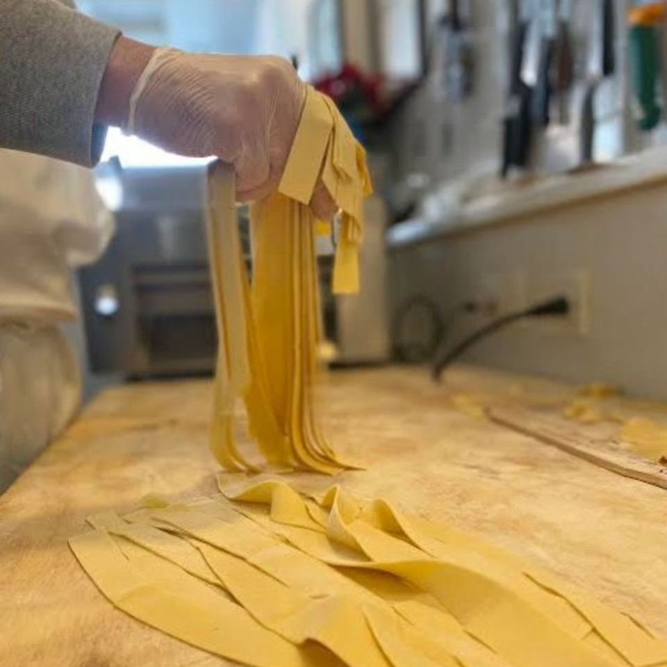 Gloved hands pull freshly cut fettuccine pasta from a machine onto a floured wooden surface.