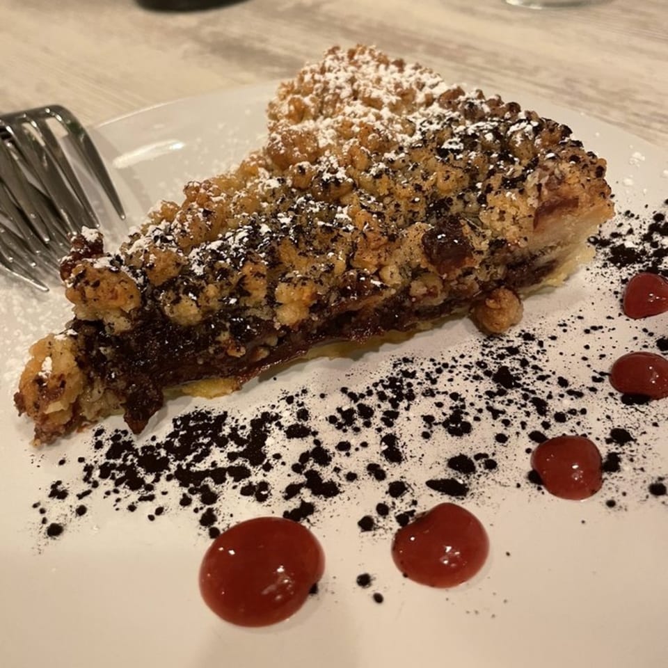 A slice of chocolate crumble cake with powdered sugar, cocoa dust, and red berry sauce.