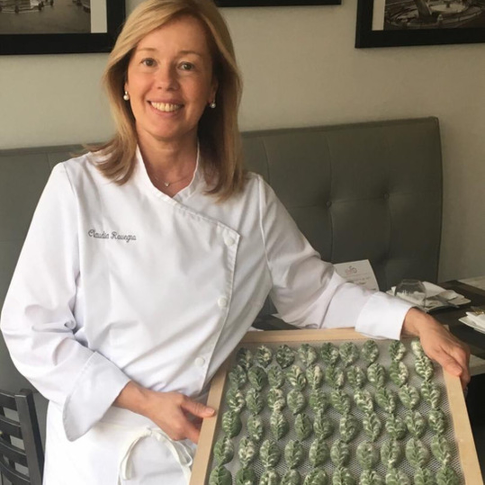 Smiling chef Claudia Ravega in a white coat holds a tray of green, leaf-shaped Italian pasta.