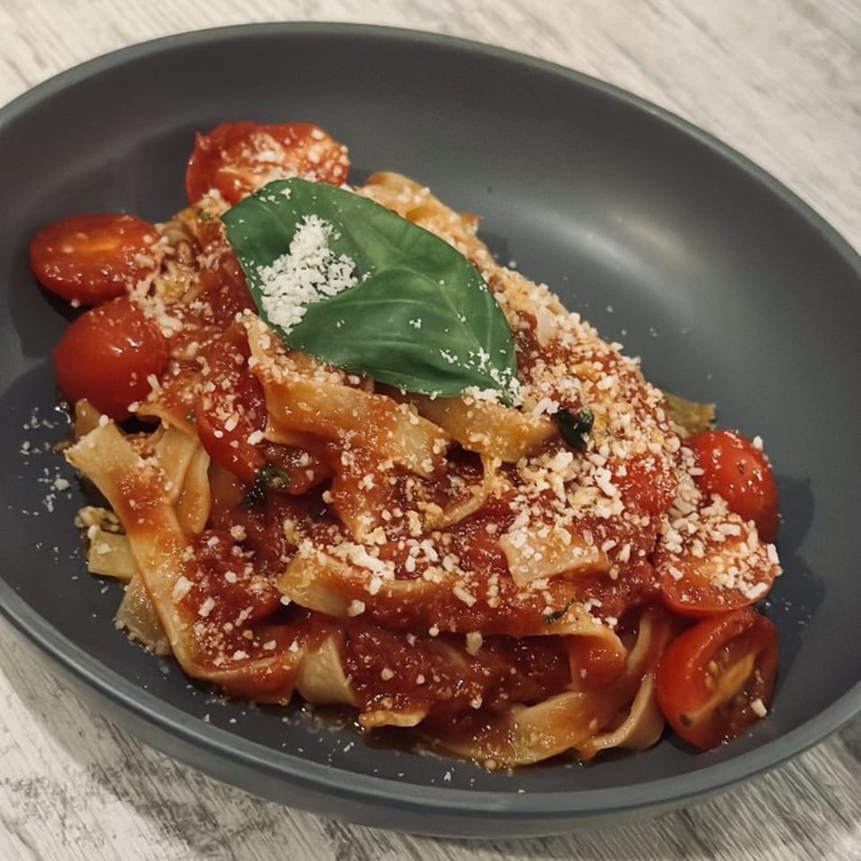 From Scratch's pappardelle pasta with tomato sauce, cherry tomatoes, parmesan, and basil in a dark bowl.
